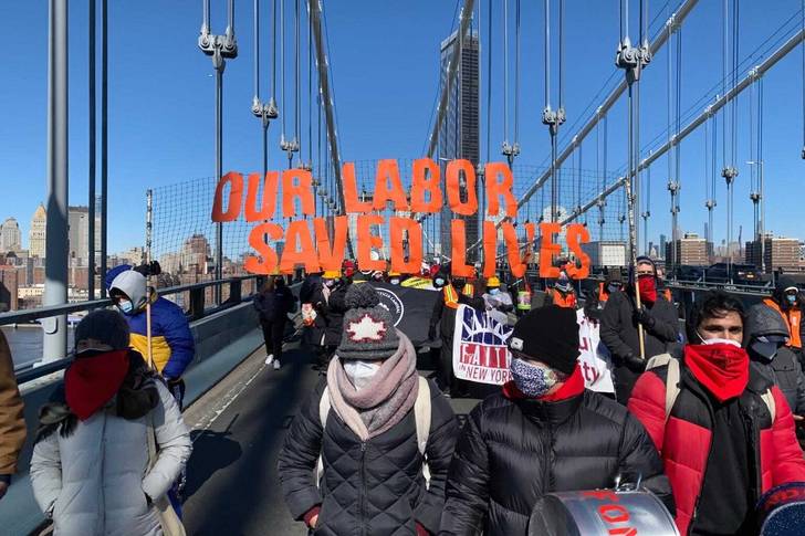 People dressed in winter coats and hats march across the Brooklyn Bridge  with orange, cutout letters above their heads that spells out "Our Labor Saved Lives."